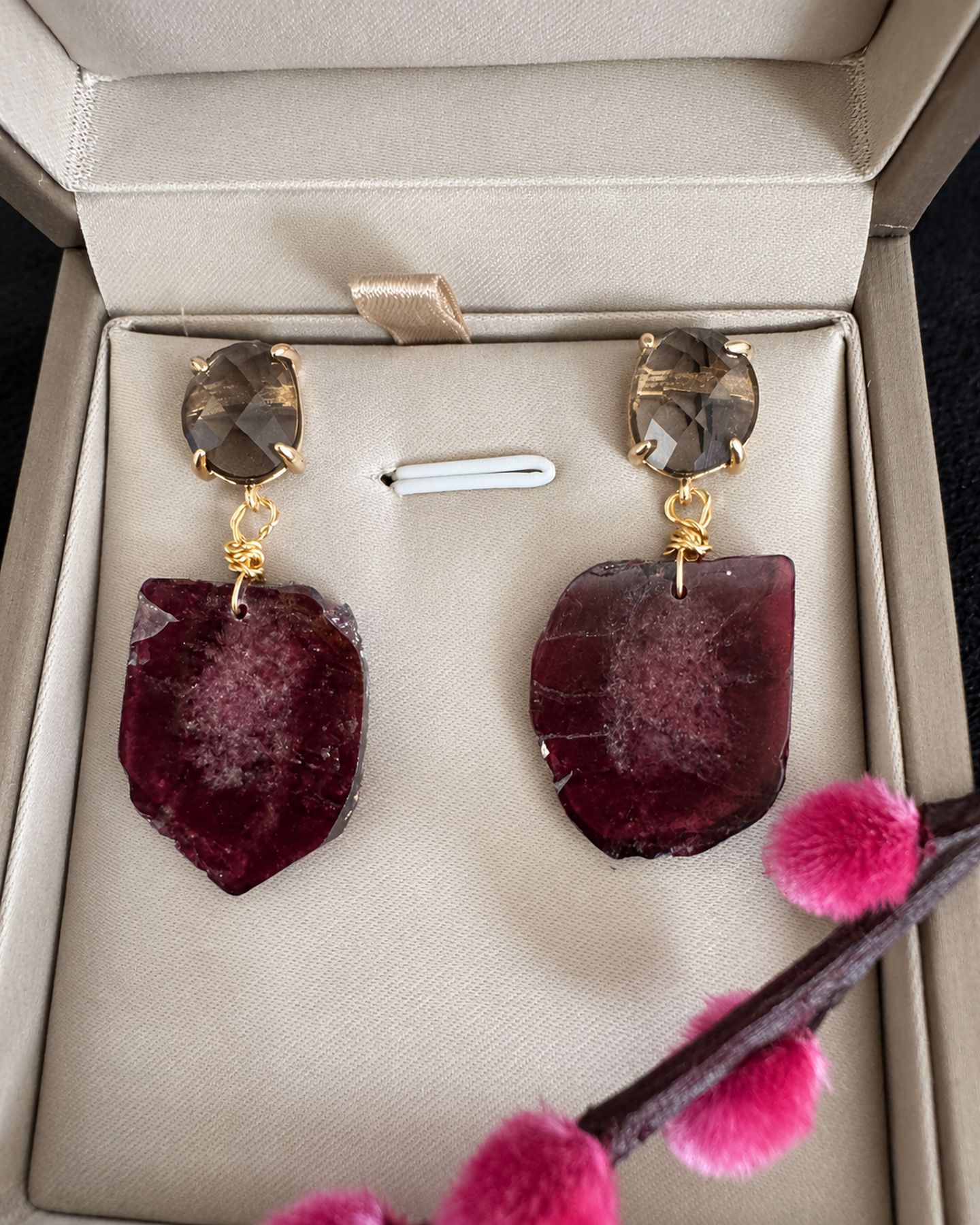 Burgundy drop earrings displayed in an open cream jewellery box with a pink flowering twig across the lower edge.