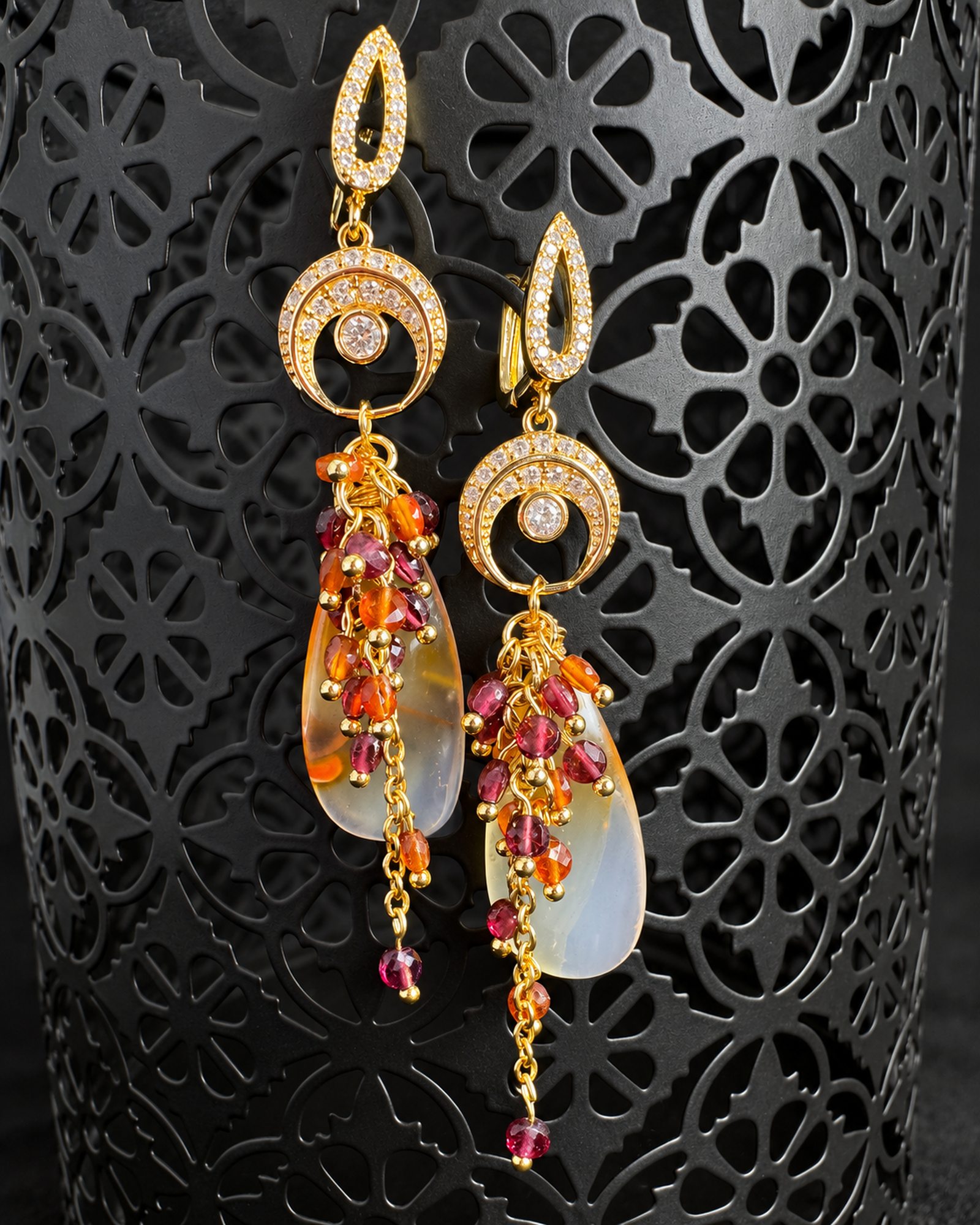 Earrings hanging at staggered heights against a dark pierced lantern, showing bead clusters, translucent drops and dangling chains.