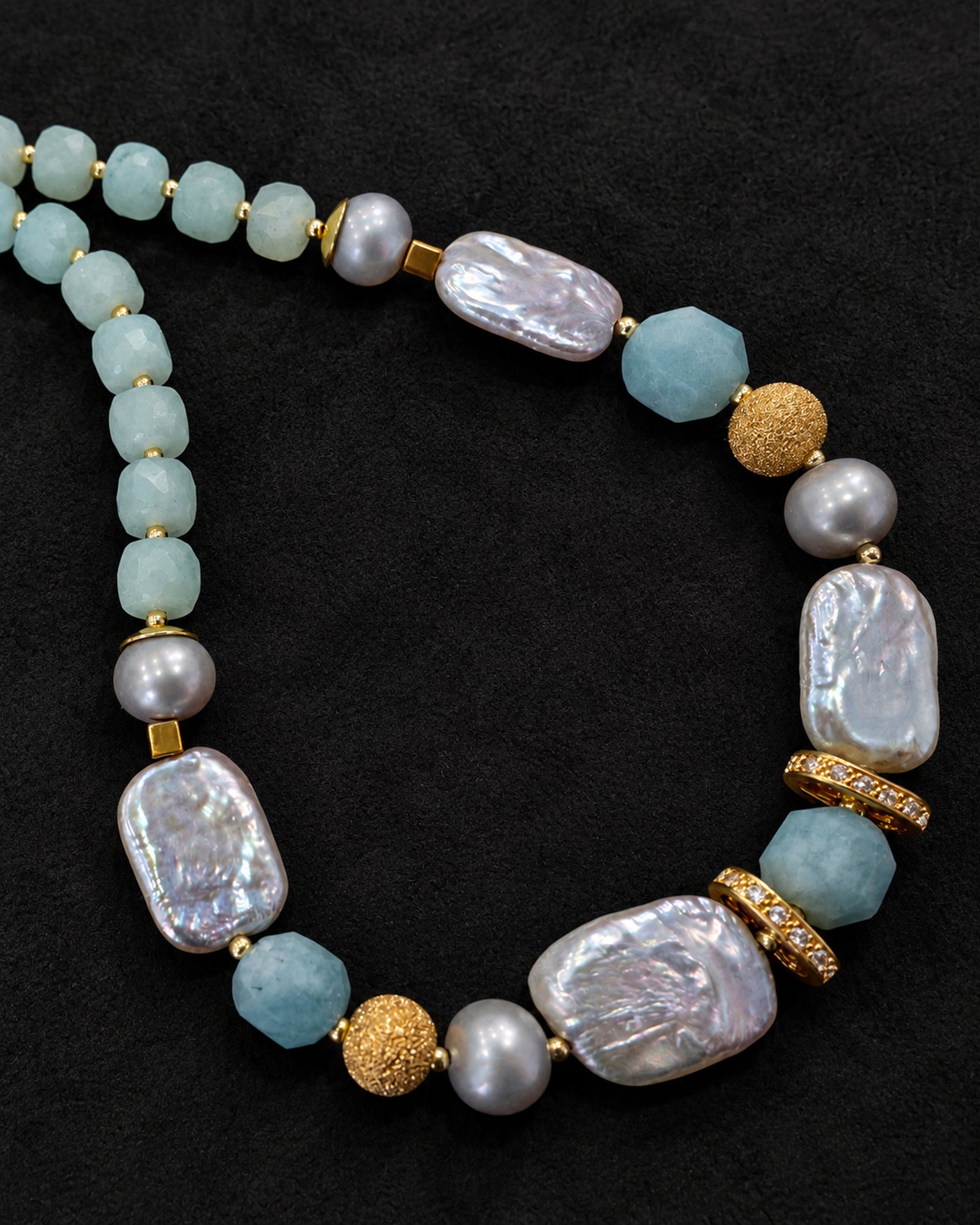 Pale aqua necklace with irregular silvery rectangular beads, rounded grey beads and gold-tone details on black fabric; upper-left strand is cropped.