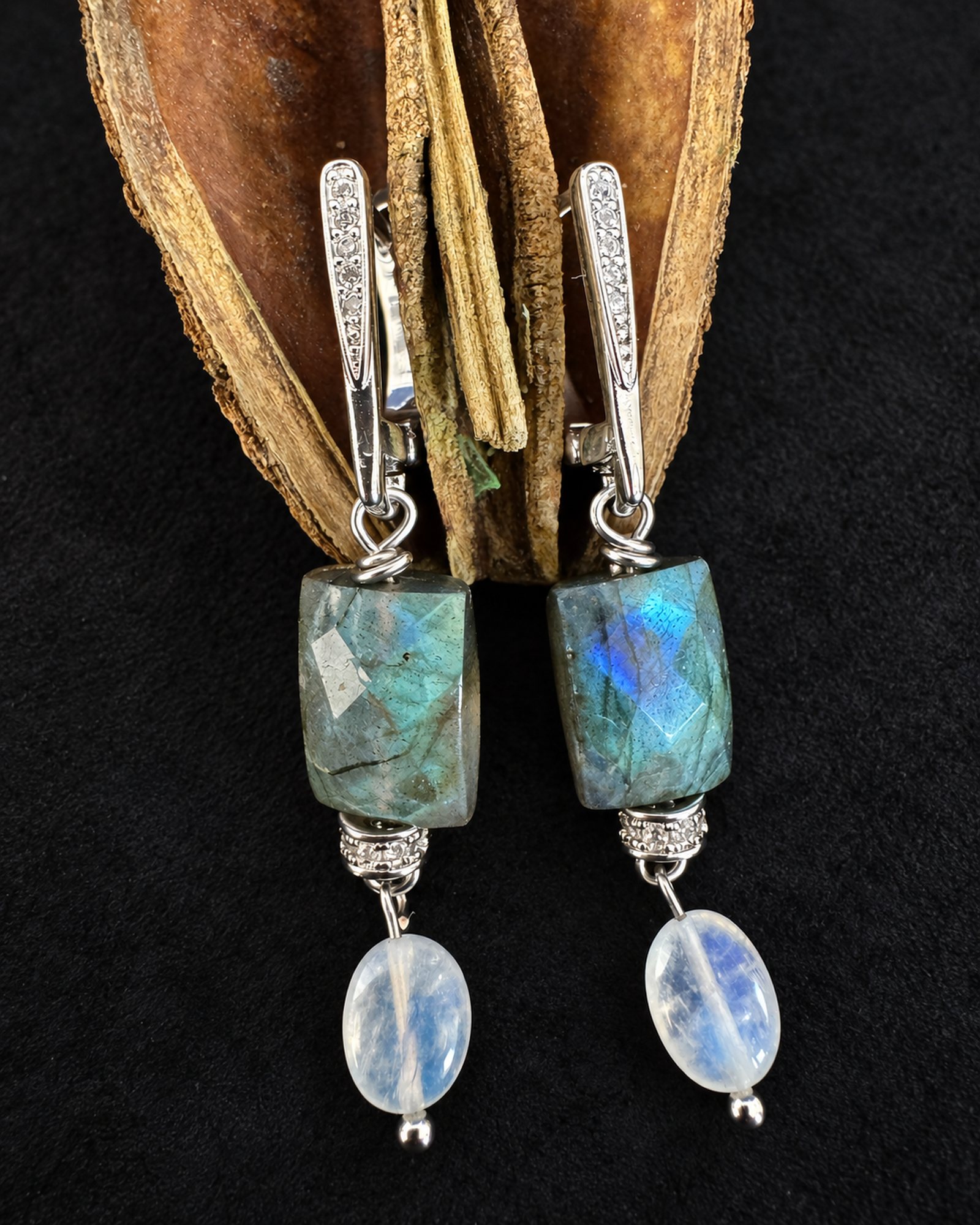 Drop earrings with mottled grey-green rectangular beads, blue reflections, pale translucent oval drops and silver-toned fastenings, hanging from a dried seedpod.