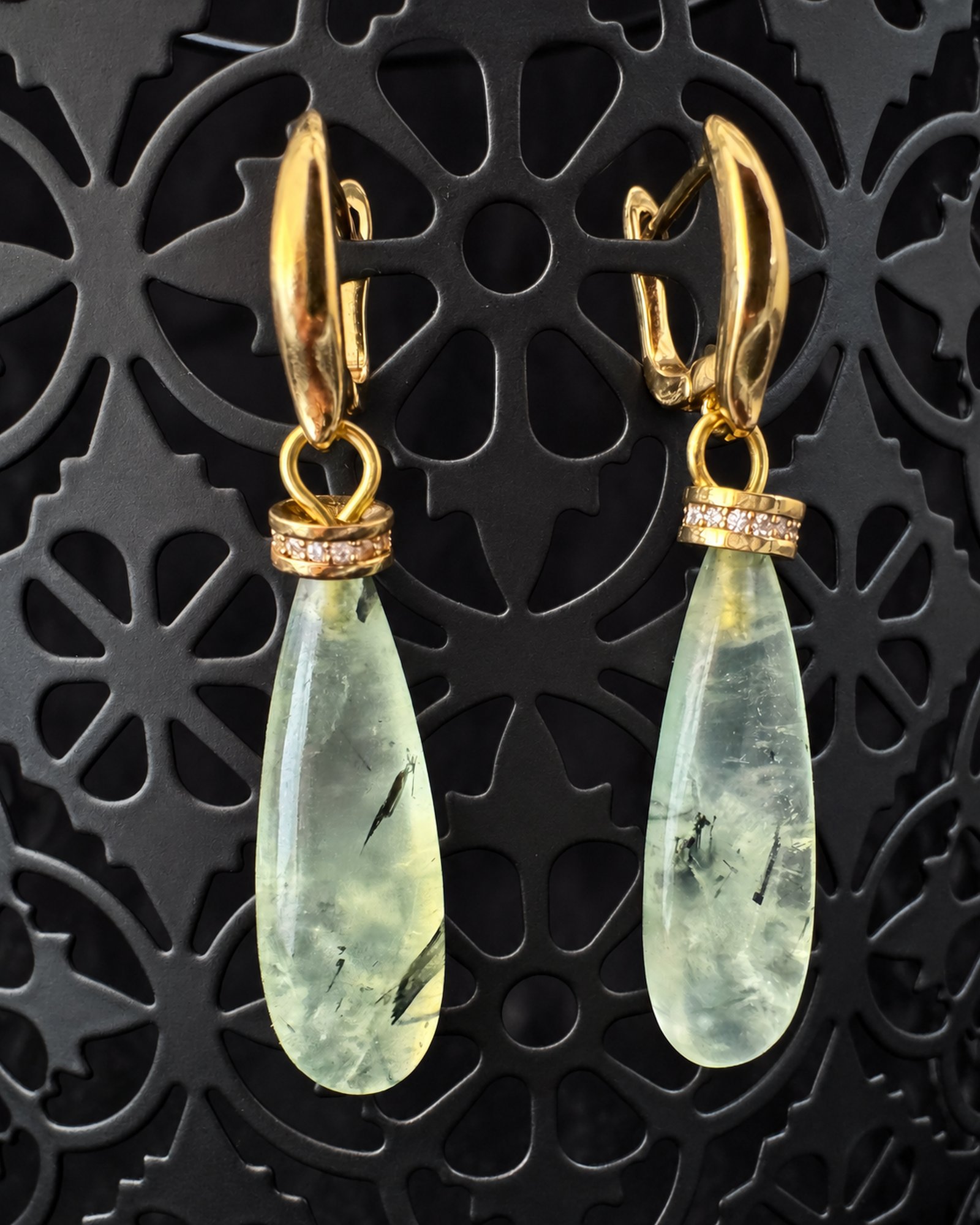 Pale green elongated teardrop earrings with dark internal markings and gold-toned fastenings hanging from a black patterned display.