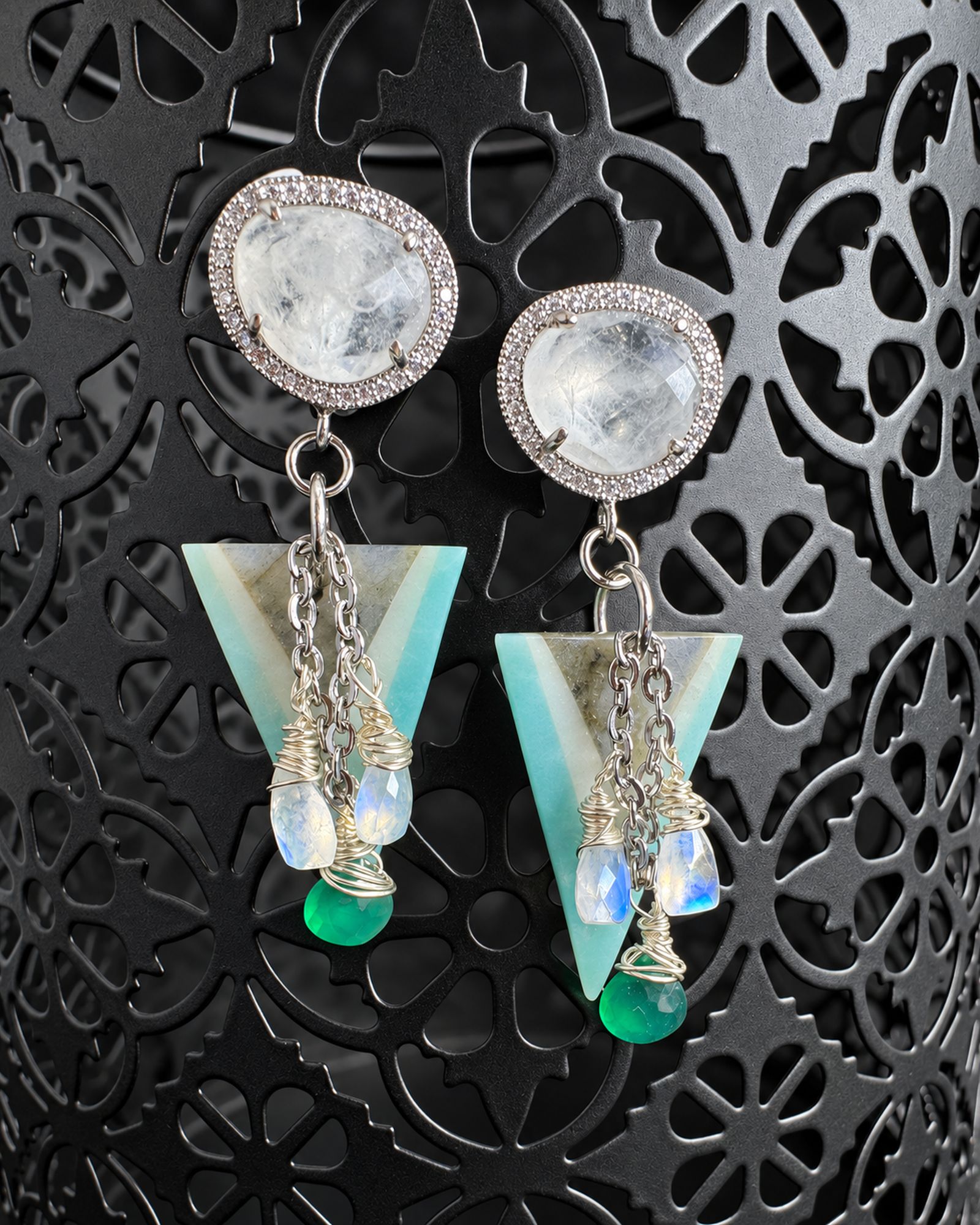 Teal Nysa earrings hanging from a dark patterned display, with pale rounded tops, aqua triangles and green drops.