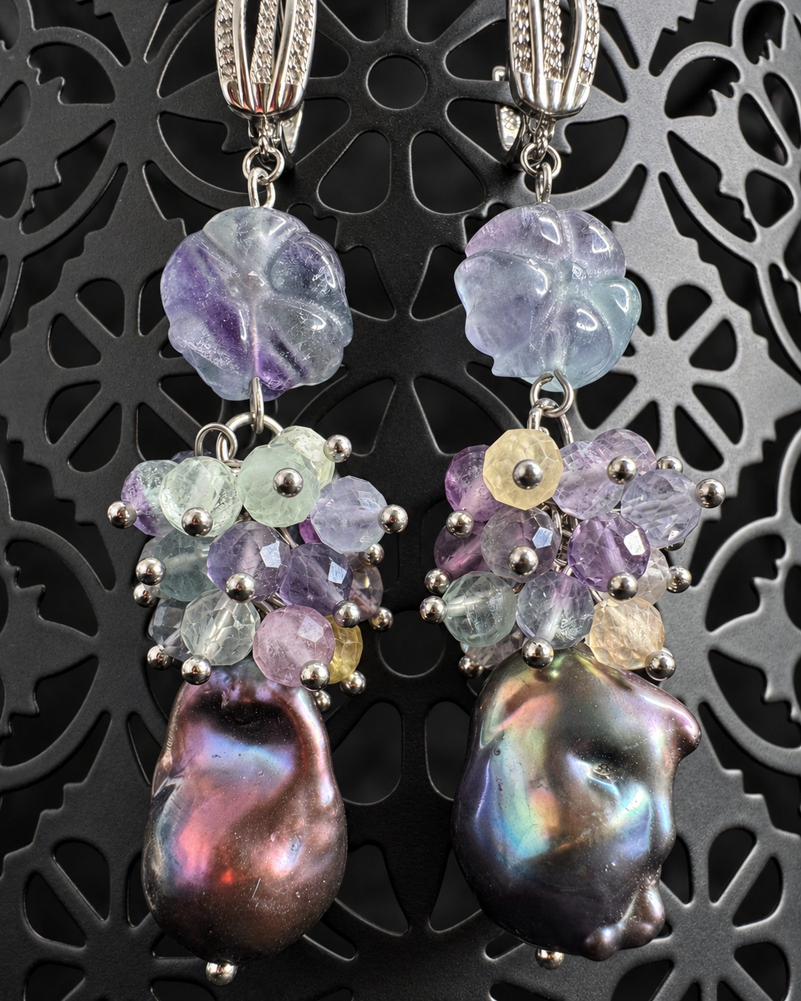 Dusk Phoebe earrings hanging against a decorative dark support, with pale bead clusters and dark iridescent drops.