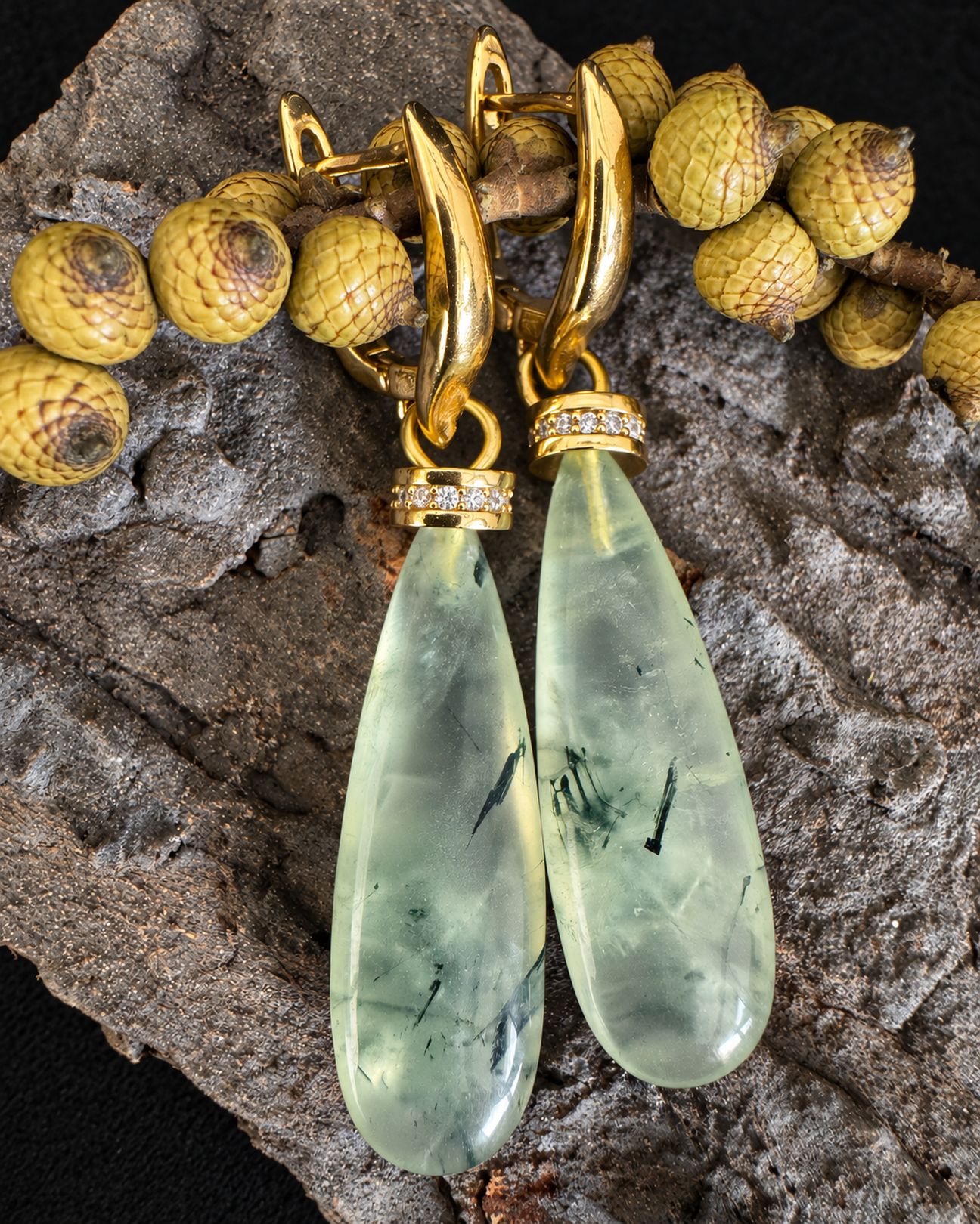 Pale green drop earrings resting against bark beneath a branch with yellow berries, with their gold-toned fastenings visible.
