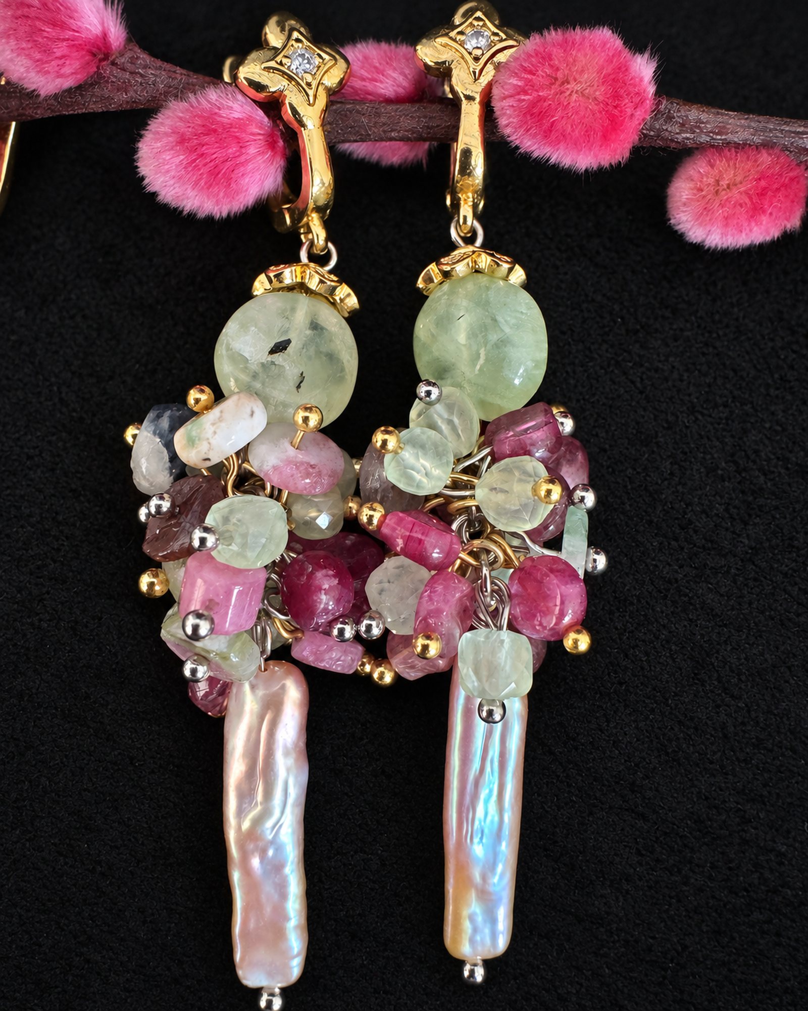 Two earrings arranged sideways on black fabric, showing pale green round beads and pink clusters beside a pink decorative branch.