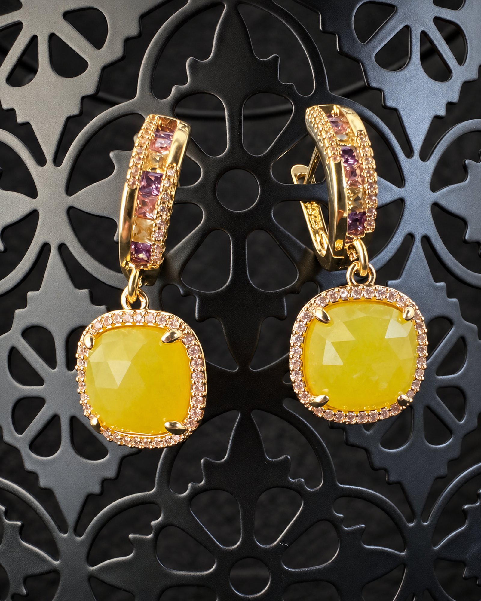 Pair of gold-toned earrings with pink-lilac hoop accents and cushion-shaped yellow drops surrounded by small clear stones, hanging against a dark ornate display.