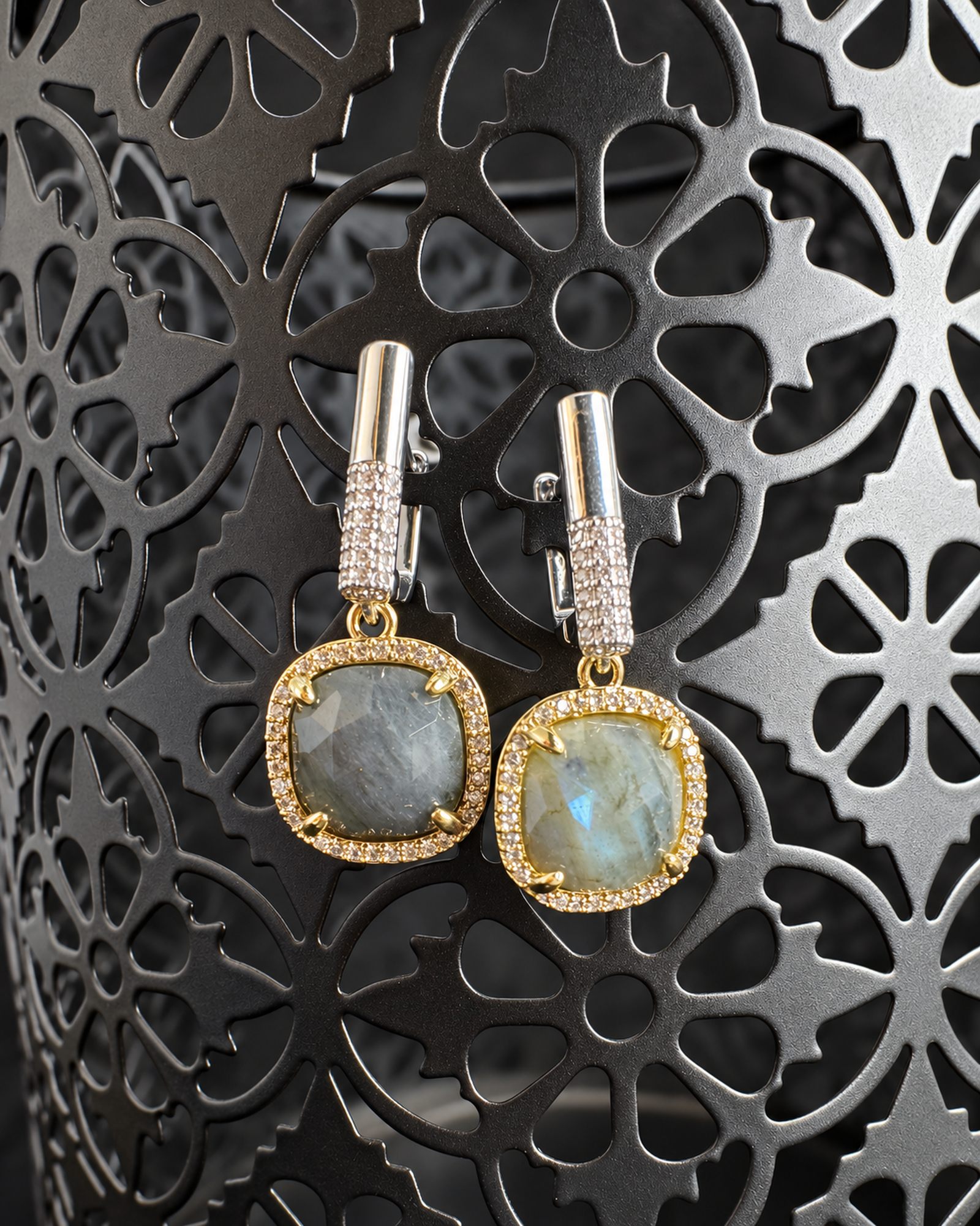 Pair of grey drop earrings hanging from a dark decorative metal stand, with a pale blue reflection in the right drop.