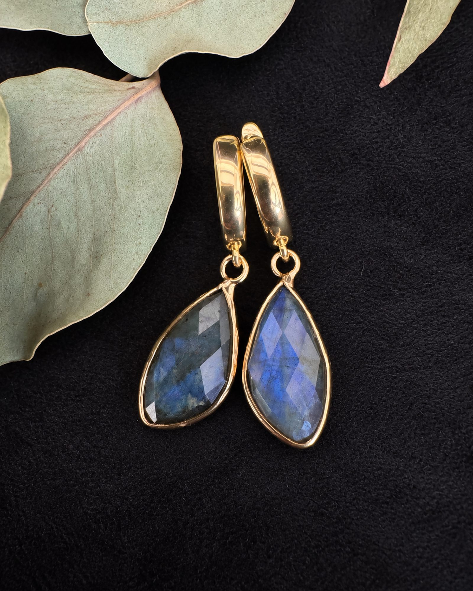 Gold-toned hoop earrings with smoky blue and violet-reflecting drops on black fabric beside pale green leaves.
