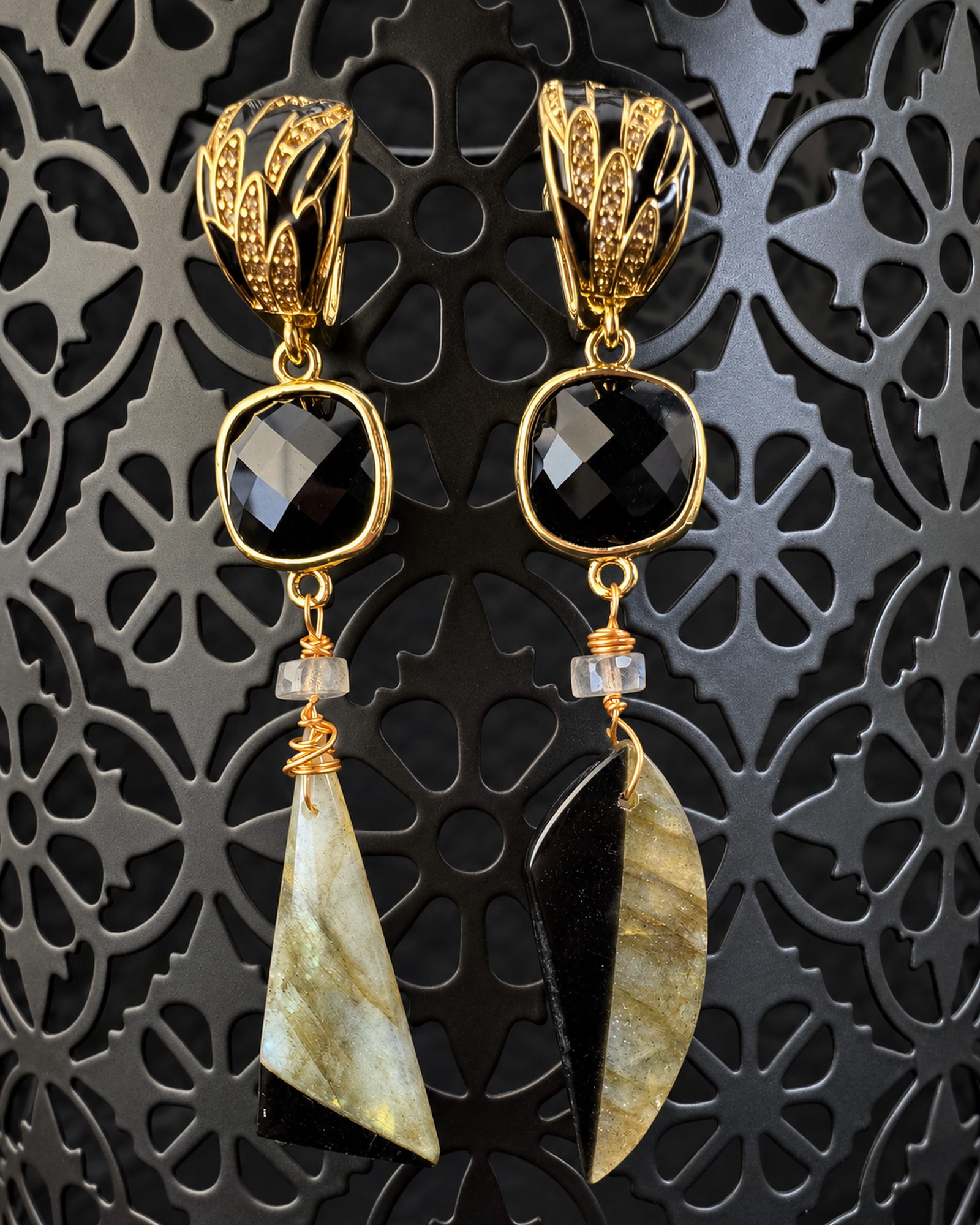Asymmetric earrings hanging on a dark patterned display, with black faceted accents, gold-toned details and contrasting triangular and curved drops.