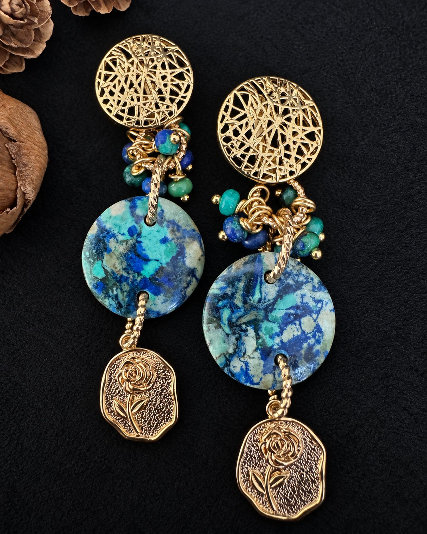 Pair of drop earrings laid flat on black, with gold-toned openwork tops, mottled blue-green round drops and rose-patterned lower charms.