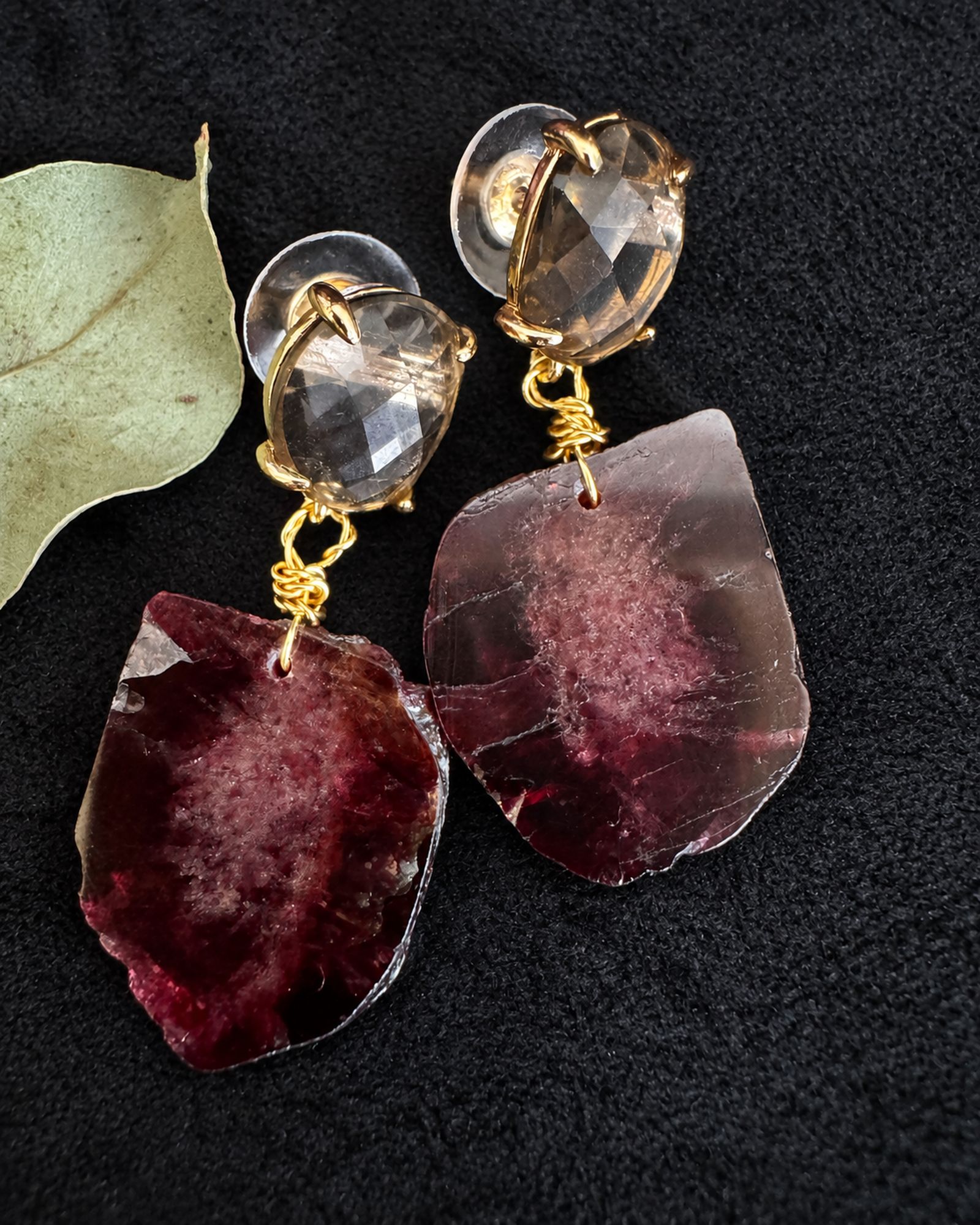 Close view of burgundy drop earrings with smoky oval tops and gold-toned connectors on a black surface beside a pale leaf.