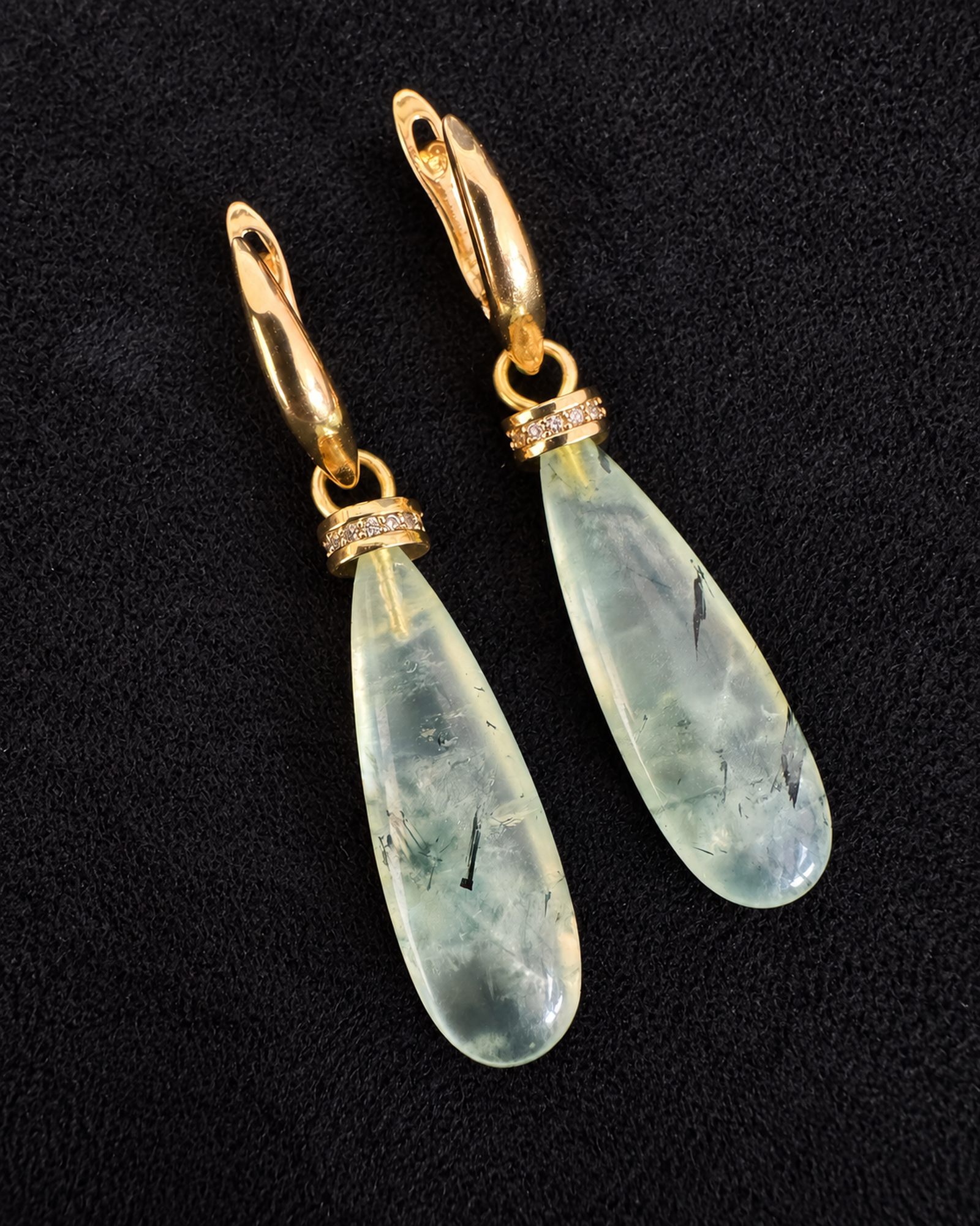 Pale green teardrop earrings arranged diagonally on black fabric, showing their gold-toned fastenings and cloudy internal markings.