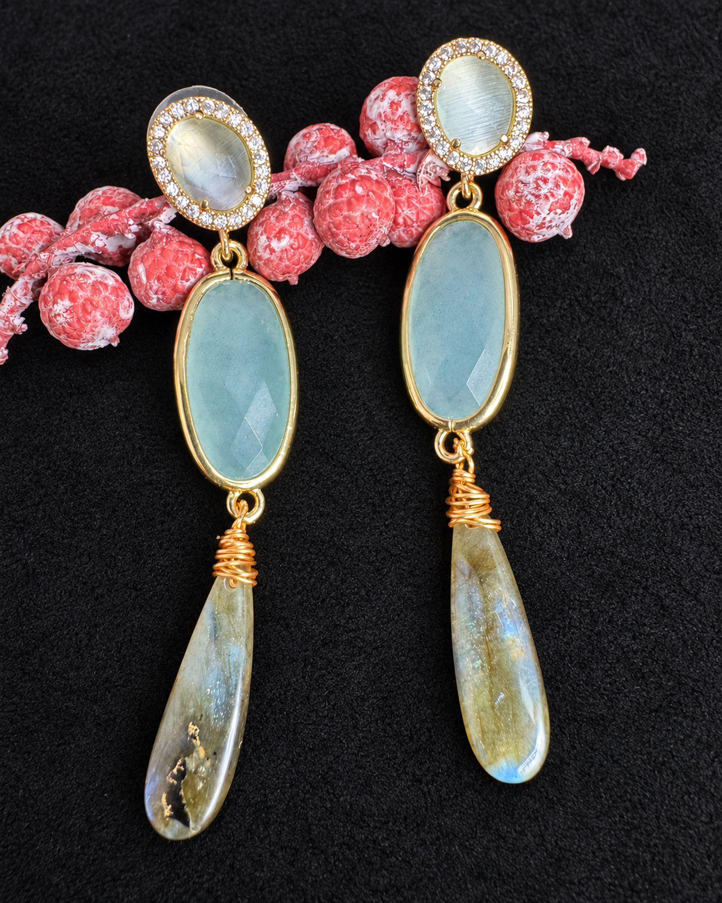 Long drop earrings with pale oval tops, clear halos, aqua oval centres and tapered smoky drops, arranged beside pink berries on black fabric.