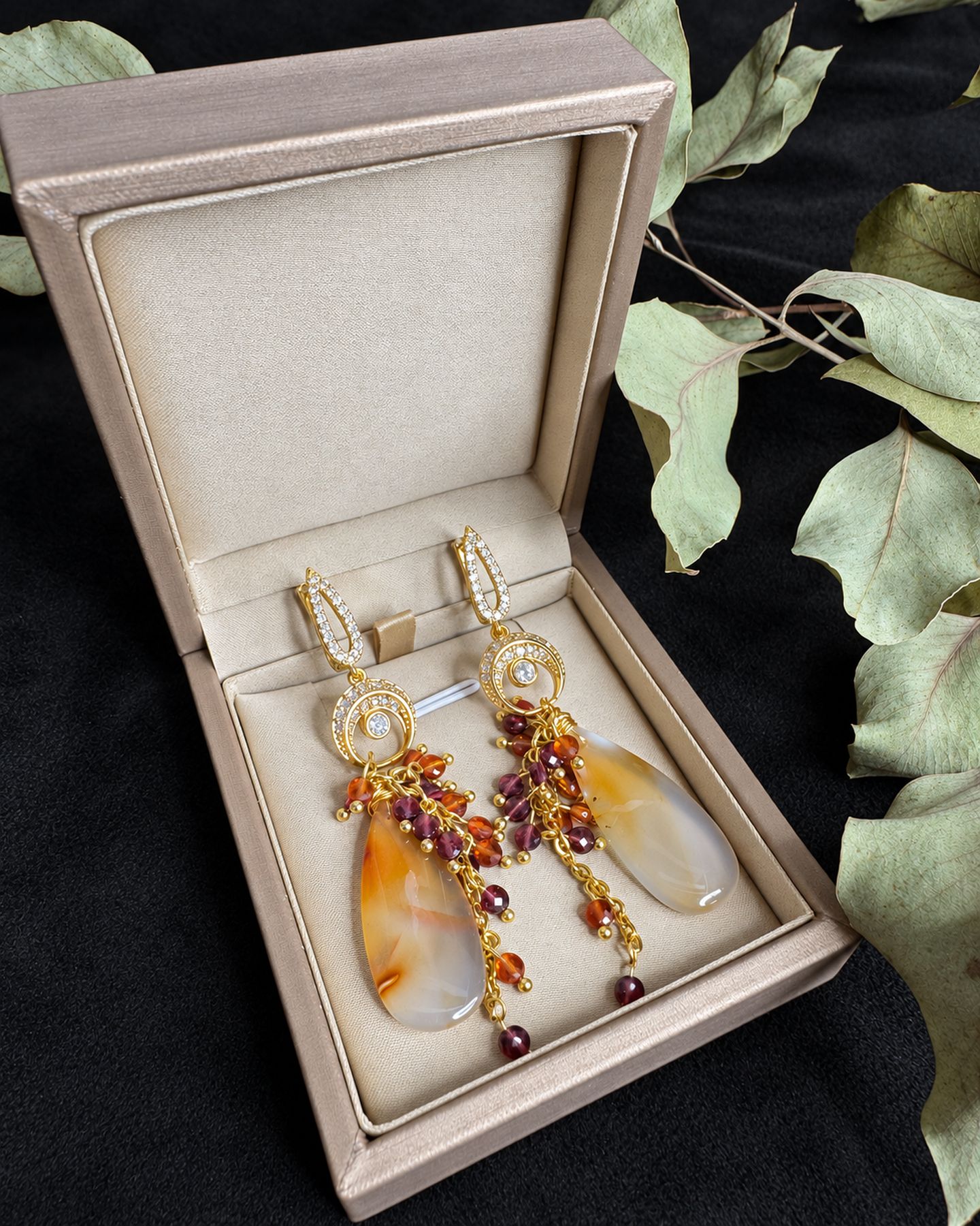 Pair of earrings resting inside an open pale jewellery box, with eucalyptus leaves beside it.