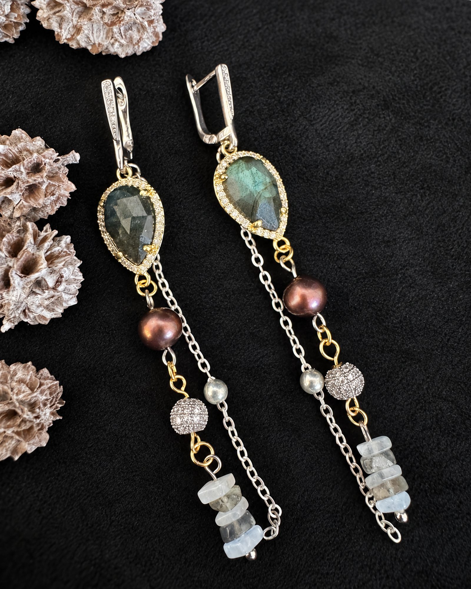 Pair of long earrings arranged on black beside pinecones, showing green pear-shaped centres, brown accents, fine chains and pale irregular drops.