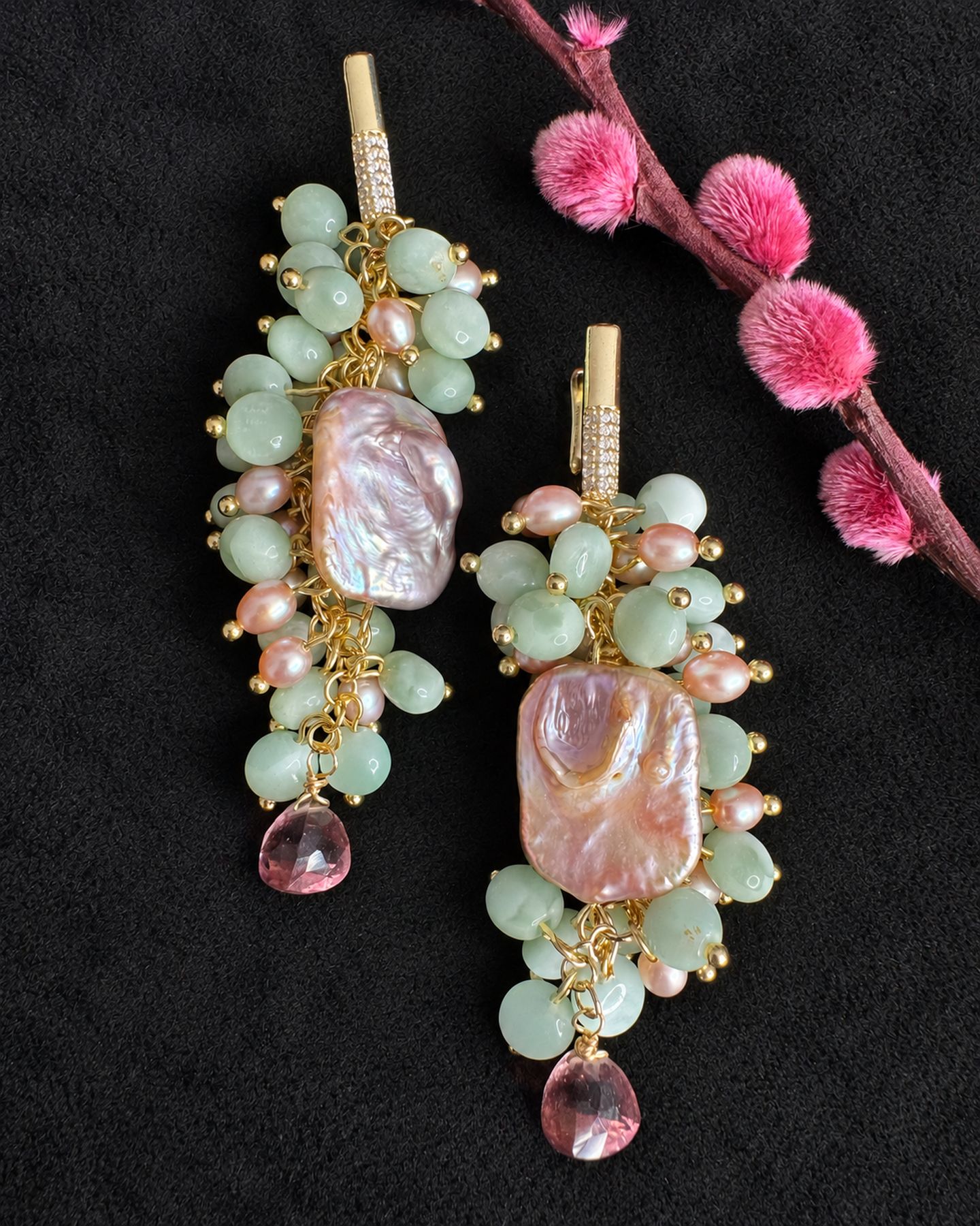 Drop earrings with pale green bead clusters, irregular pink-lavender centres and purple tips, arranged diagonally on black beside a pink branch.