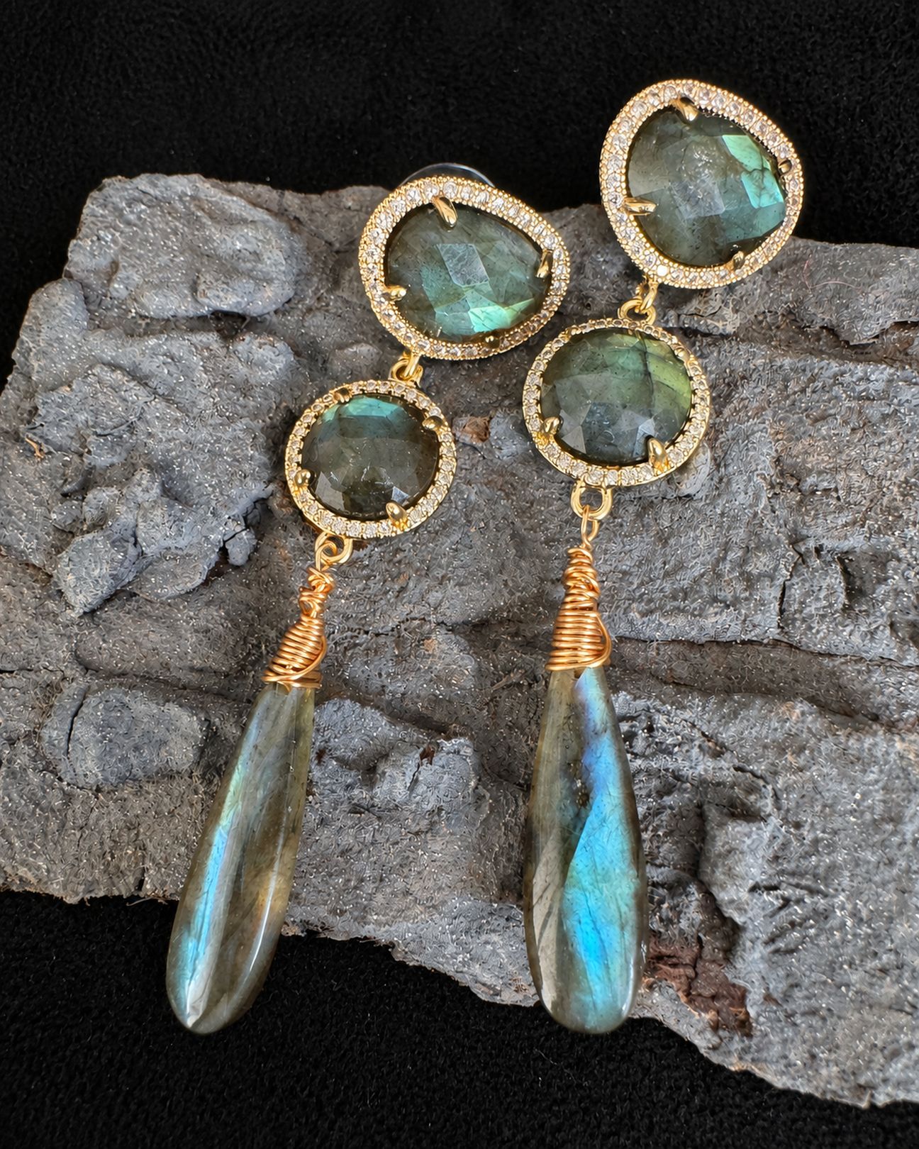 Horizontal close-up of the earrings on grey bark, showing round green-grey stones, wrapped gold-toned wire and blue reflections in the drops.