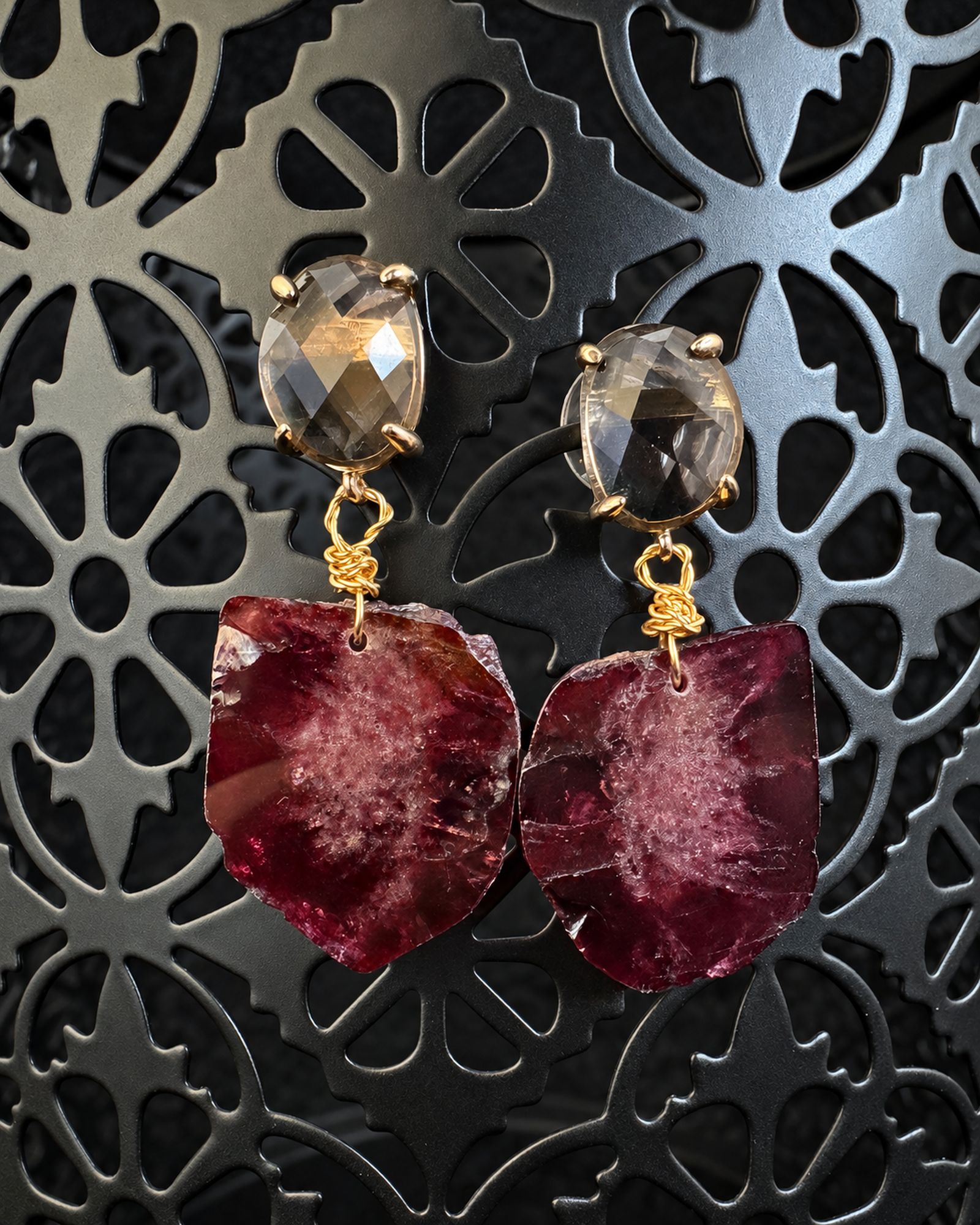 Pair of earrings with smoky oval faceted tops and irregular burgundy drops hanging from a dark ornate display.