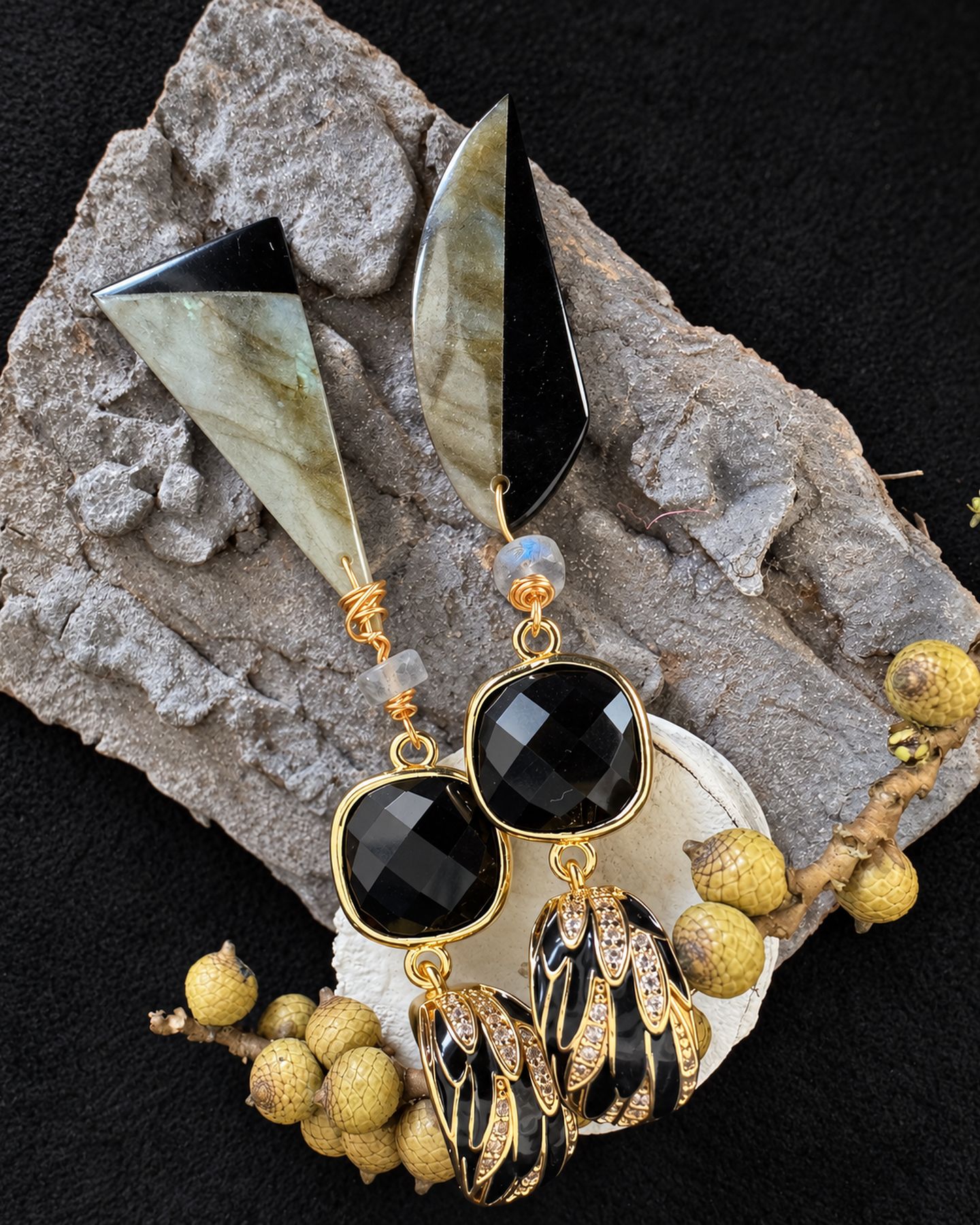 Asymmetric earrings arranged diagonally on grey bark beside a pale shell and yellow botanical sprig.
