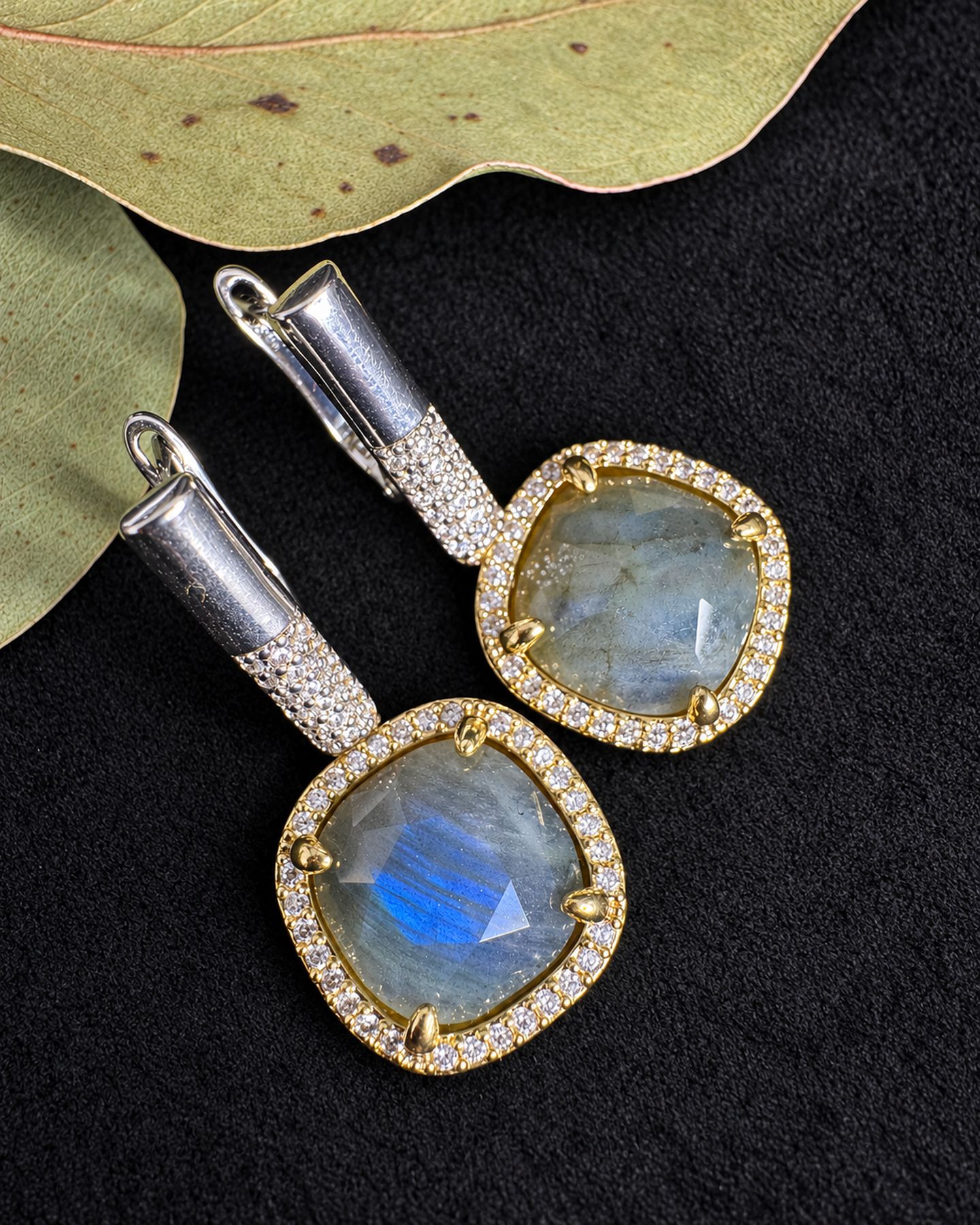 Earrings arranged diagonally on black fabric beside leaves, showing a blue reflection in the lower grey drop.