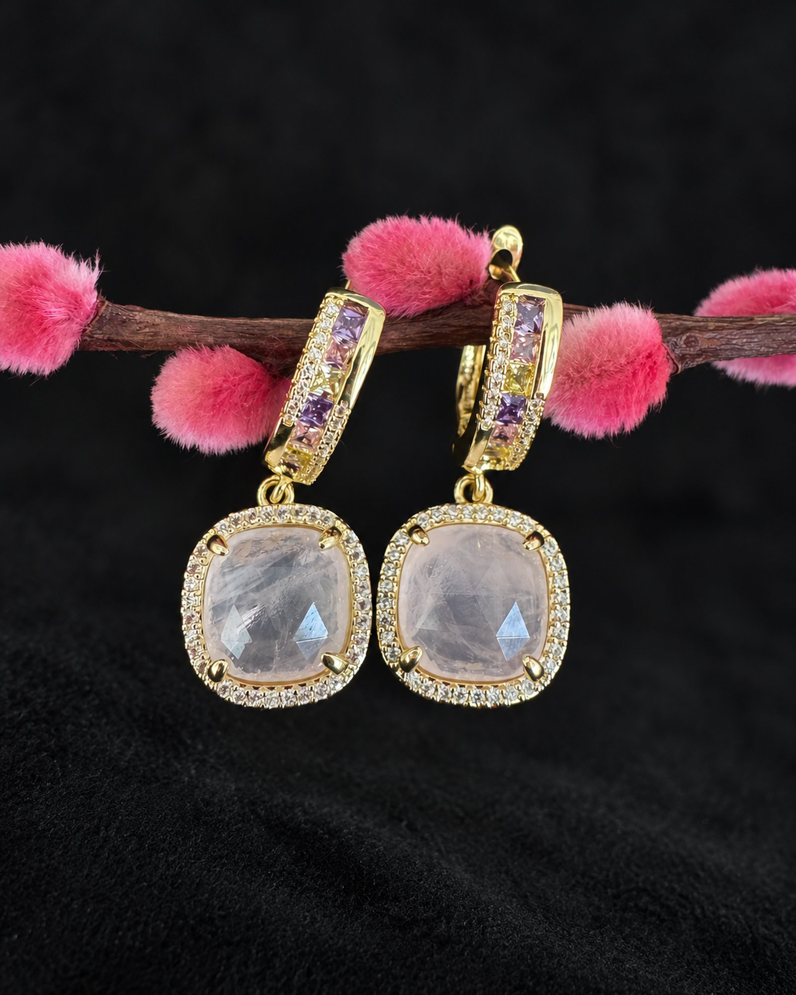 Pair of gold-toned earrings with pale smoky lilac cushion-shaped drops, hanging from a branch with pink buds.