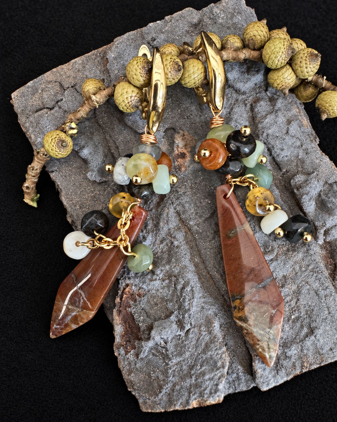 Pair of long earrings with reddish-brown pointed drops and multicoloured bead clusters arranged on a grey slab beside seed pods.