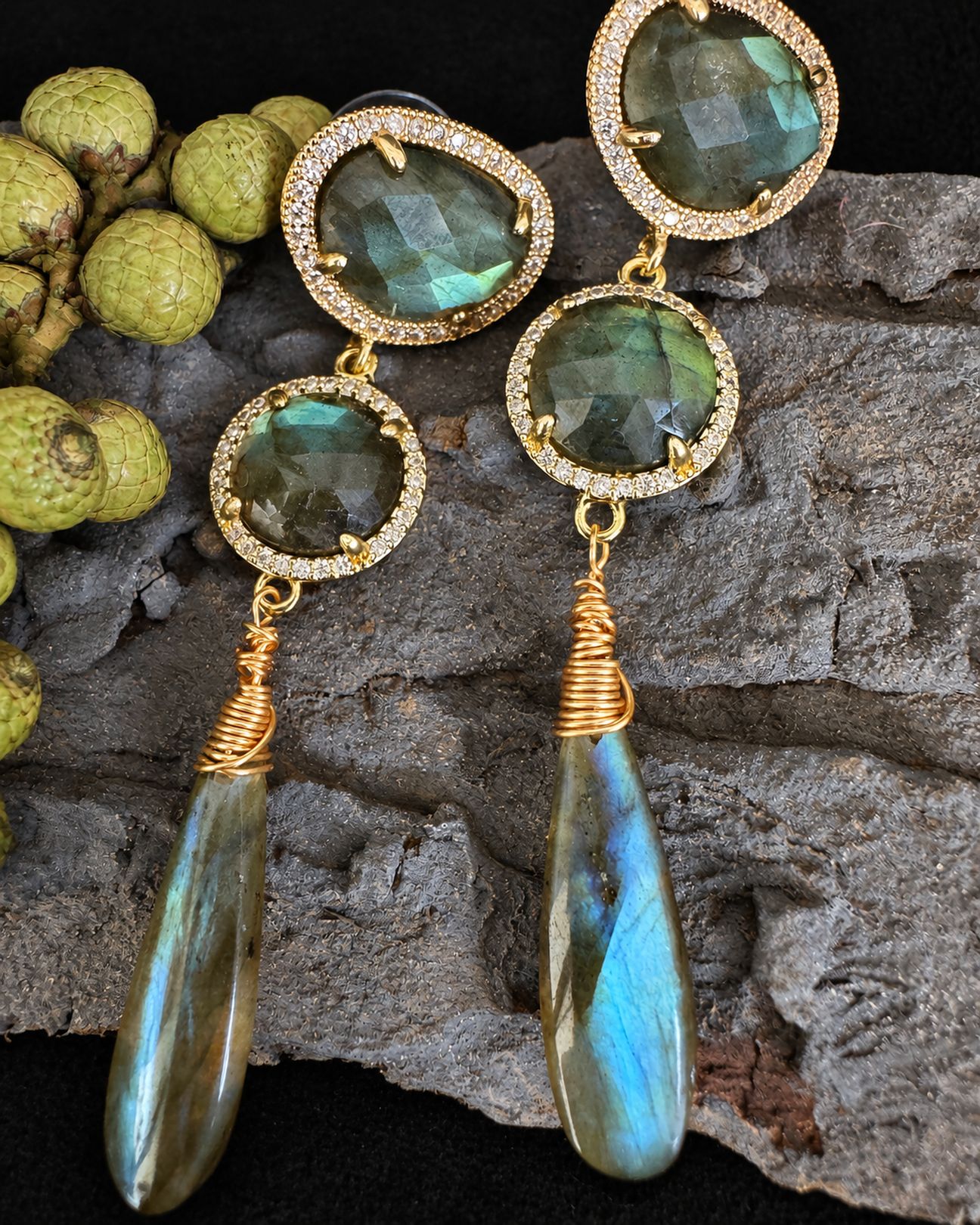 Earrings arranged vertically on grey bark beside green berries, with smoky green stones and a bright blue reflection in the right drop.
