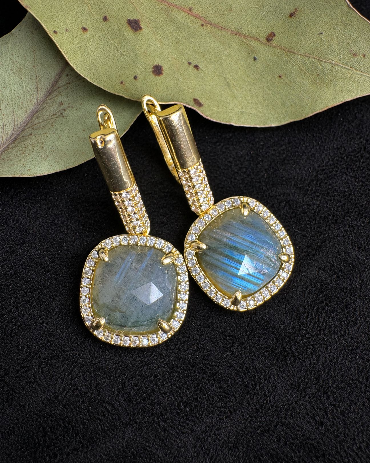 Gold-toned drop earrings with cushion-shaped grey centres, blue reflections and clear halos, resting on black fabric beside leaves.