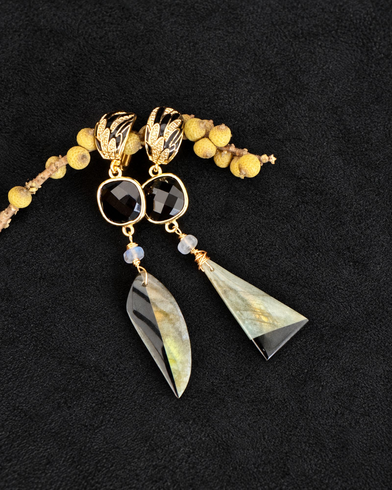 Full pair of asymmetric earrings arranged on black fabric beside a yellow botanical branch.