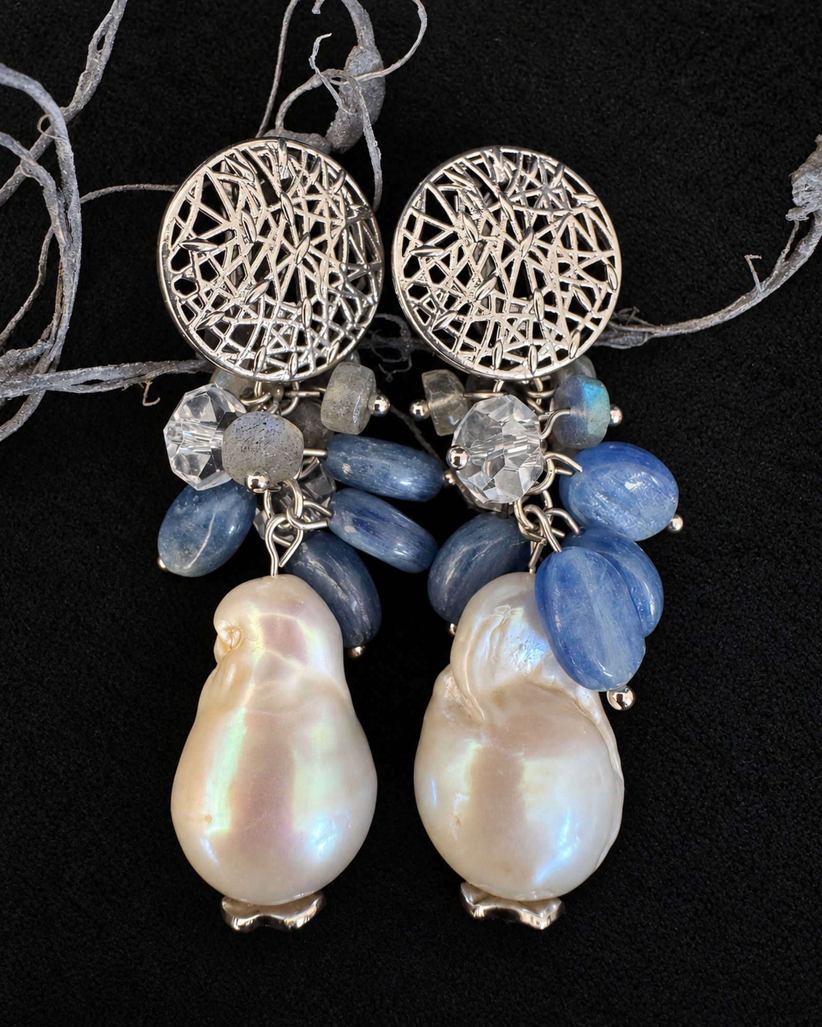 Front flat lay of earrings with silver-toned openwork discs, blue bead clusters and irregular ivory-coloured drops on black fabric.